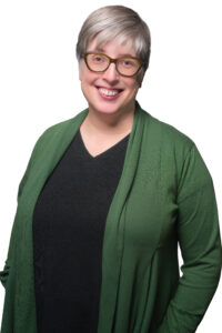 Headshot of a smiling white woman with glasses and greying hair, wearing a green cardigan over a black V-neck.