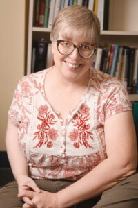 A white woman with glasses and short, greying hair sits on the floor in front of a bookcase, smiling