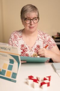 A white woman with glasses and short, greying hair sits at a table holding a clipboard and pen. In front of her are some brightly-colored red and white blocks and a propped-up test manual.